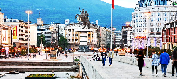 Panoramic view of Skopje with the stone bridge at sunset and  people walking across it. At the back is seen Macedonia Square with its large statue of Alexander the Great on the center.