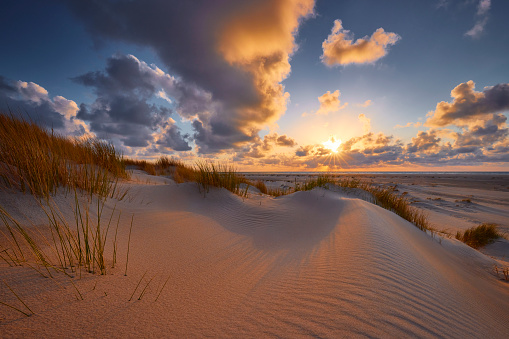 Sand dunes during sunrise. There is a beautiful pattern of lines formed in the sand. The sun shines brightly through the clouds and the rest of the sky is blue. There is grass on the dunes.