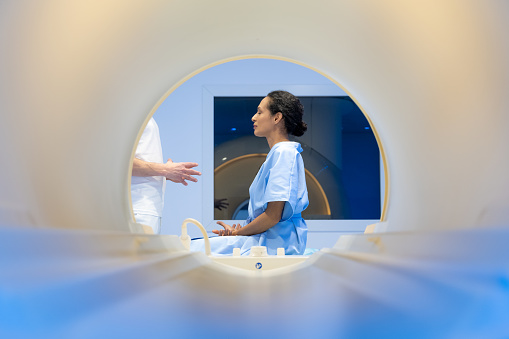 Mature woman in hospital gown sitting on the MRI examination bed and talking with a male doctor in hospital.
