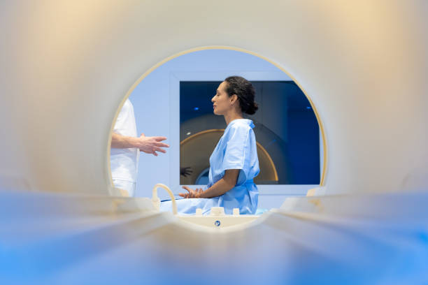Mature woman talking with a doctor before MRI scan Mature woman in hospital gown sitting on the MRI examination bed and talking with a male doctor in hospital. oncology stock pictures, royalty-free photos & images