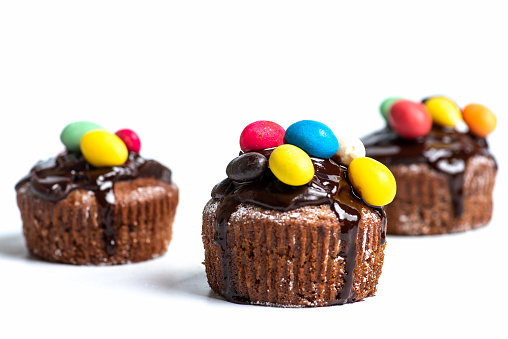 Chocolate muffins or brownies in a paper cupcake with chocolate cream and colorful candy bonbons on white background isolated