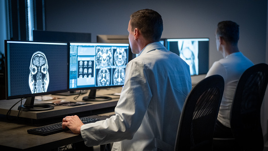 Rear view of male radiologist looking at the MRI image of the head on his monitor and analysing it.