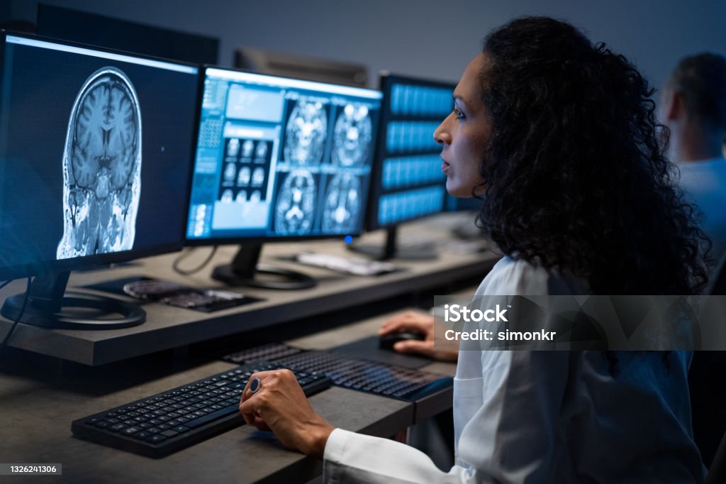Female radiologist analysing the MRI image of the head Side view of female radiologist looking at the MRI image of the head on her monitor and analysing it. MRI Scan Stock Photo