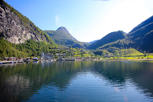 Beautiful view of Geiranger town from the water on a summer day. Reflections of the mountains in the fjord. lush green landscape with peaks towering over the village. Norway.