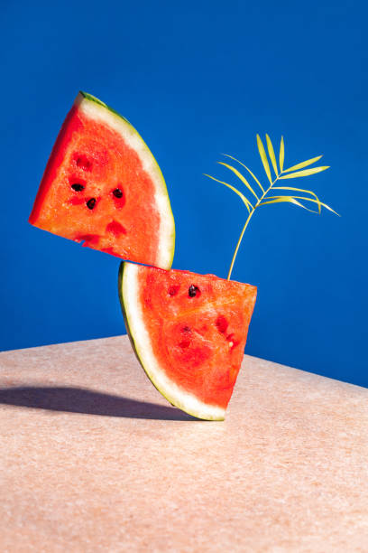 istock Two pieces of watermelon with a palm leaf balancing on a blue background 1326244909