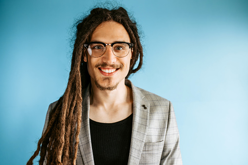 Portrait of a young modern stylish man with dreadlocks hairstyle, wearing a eyeglasses and suit, standing against blue background, smiling, looking at camera.