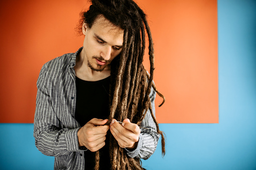 Portrait of young handsome stylish man with dreadlocks hairstyle, taking care of his hair, standing against blue - orange background.