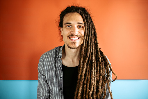 Portrait of a young modern stylish man with dreadlocks hairstyle, wearing a shirt, standing against blue - orange background, smiling, looking at camera.