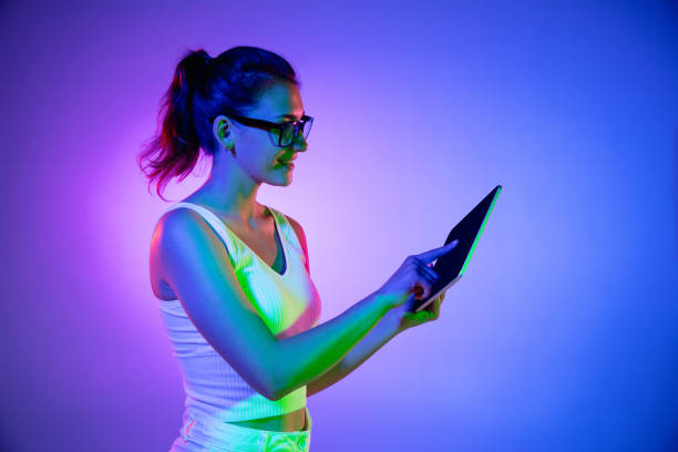 istock Holding tablet, crazy excited. Caucasian young woman's portrait on black studio background in neon light. Beautiful male model in white shirt. 1326262921