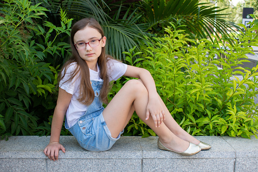 A girl sits on a granite curb and looks at us.  The girl with glasses is dressed in a white T-shirt and denim overalls.  A girl with fair skin and dark hair.