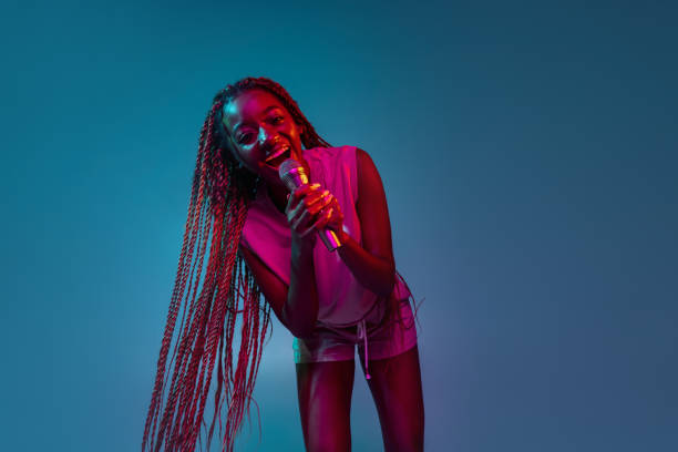 portrait d’une jeune femme africaine charmante avec micro isolé sur fond bleu dans des néons. concept d’expression faciale et d’émotion humaine. - black celebration colors dark photos et images de collection