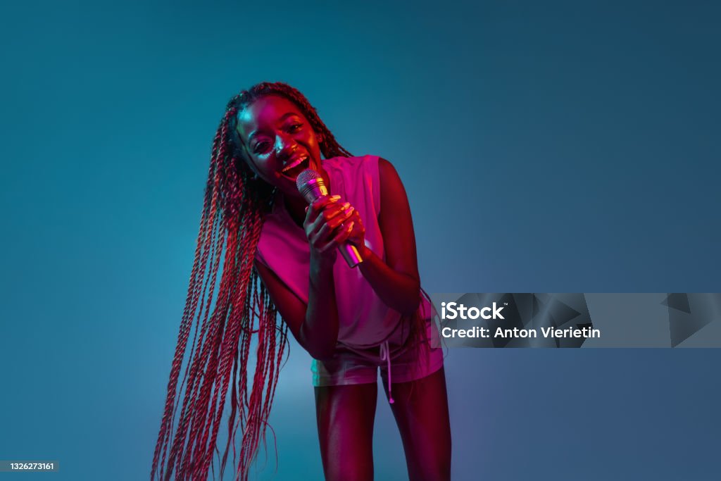 Portrait of young charming african woman with microphone isolated over blue background in neon lights. Concept of facial expression and human emotion. Singing, performing. Young joyful smiling artistic african woman singing with microphone isolated over blue background in neon lights. Concept of facial expression and human emotion. Singer Stock Photo