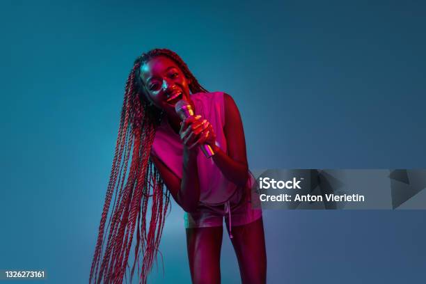 Portrait Of Young Charming African Woman With Microphone Isolated Over Blue Background In Neon Lights Concept Of Facial Expression And Human Emotion Stock Photo - Download Image Now