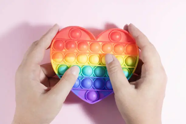 Photo of Colorful Antistress Pop it toy. Rainbow sensory fidget isolated on pink background, in the hands during the game. New trendy silicone toy. Toy for the development of fine motor skills of the fingers.
