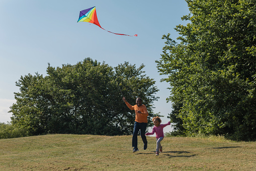 A grandad and his young granddaughter running down a hill on a bright sunny summers day flying their multicoloured rainbow kite down together. They both have big smiles on their faces and are having fun together.