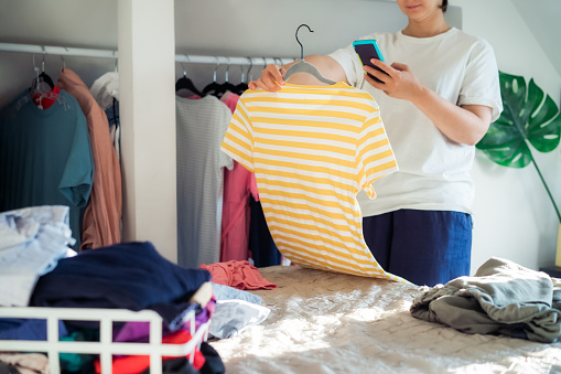 Mujer tomando foto de la camiseta usando su teléfono inteligente para vender o donar su ropa. Despejar la ropa, clasificar y limpiar. Reutilización, concepto de segunda mano. Consumidor consciente photo