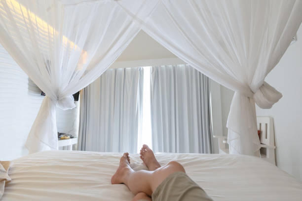 Man stretching legs in mosquito net on the bed at vacation, Sunlight through white curtain in bedroom