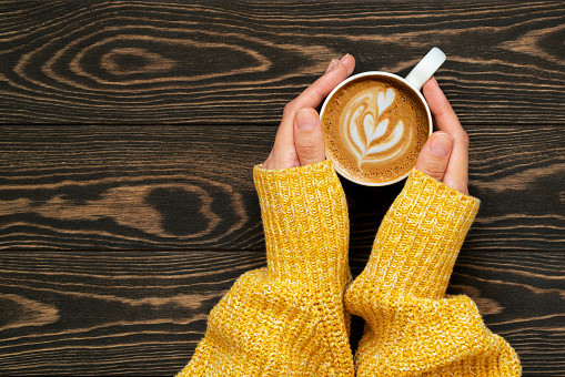 Woman hands in yellow sweater holding cup of fresh coffee with beautiful latte art on wooden background. Cozy autumn morning cappuccino closeup. Coffee break. Copy space, top view. Hygge concept.
