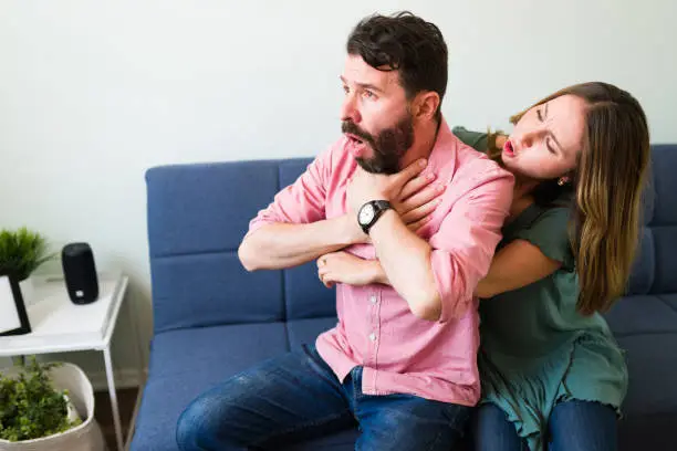Beautiful young woman telling her boyfriend to calm down while doing the Heimlich maneuver in the living room