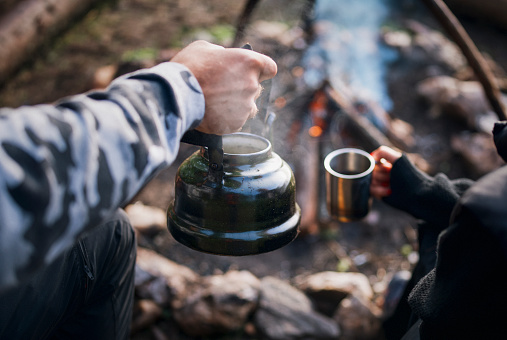 Unrecognizable couple is preparing tea in vintage teapot around campfire in the morning.