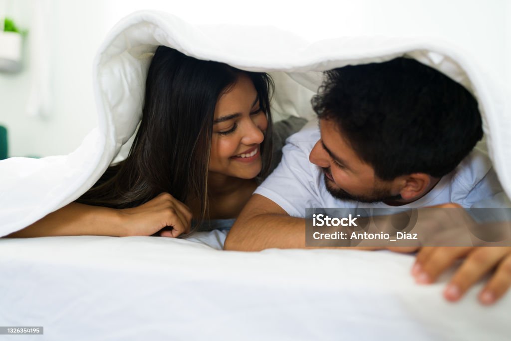 Beautiful couple hiding under the blankets Playful latin boyfriend and girlfriend lying in bed under the covers. Attractive couple enjoying their company on a leisure day Couple - Relationship Stock Photo