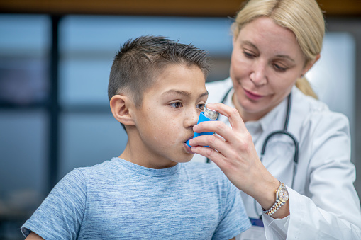 A boy sits on the examination table breathes into an inhaler that his doctor is holding.