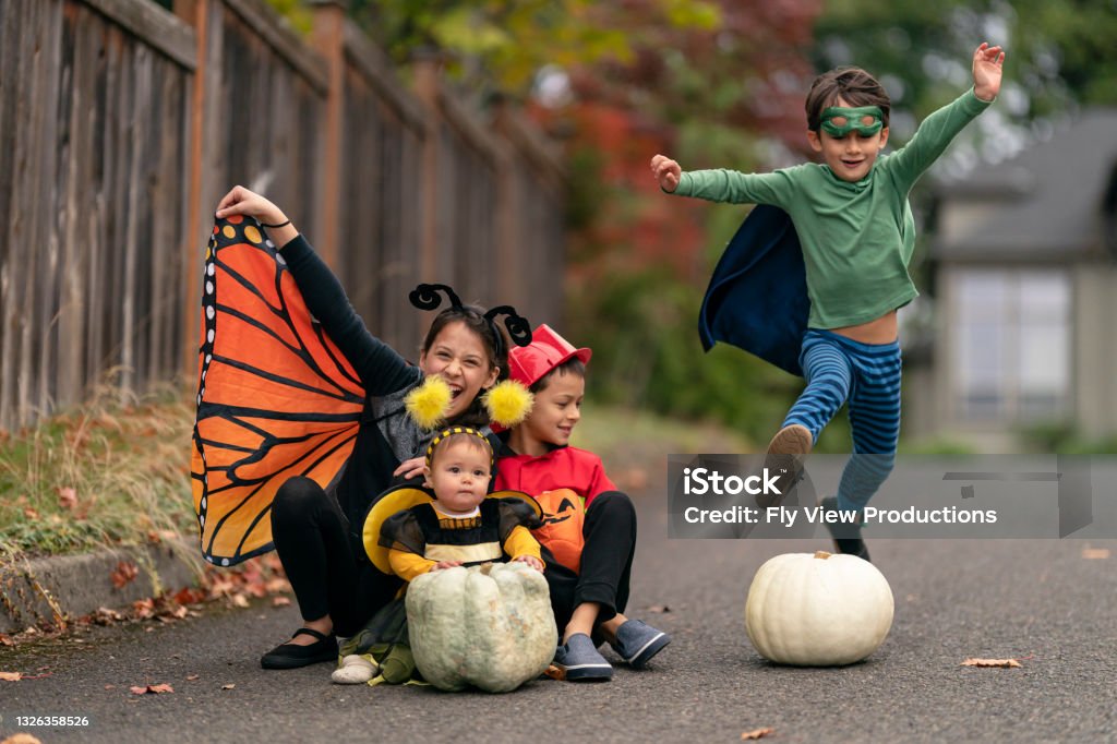Happy group of kids on Halloween Four mixed race siblings in costumes happily play together outside in their driveway on Halloween. An elementary age boy dressed as a superhero is jumping over a large white pumpkin. Halloween Stock Photo