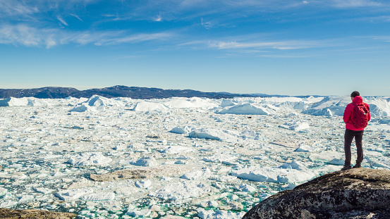 Travel in arctic landscape nature with icebergs - Greenland tourist man explorer - tourist person looking at amazing view of Greenland icefjord - aerial video. Man by ice and iceberg in Ilulissat.