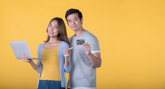 Asian couple holding credit card and laptop while looking at a camera isolated on yellow background