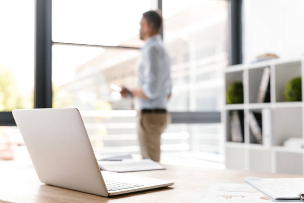 istock Image of workplace with open silver laptop lying on desk while defocused caucasian man standing and looking through big window on background 1326385610