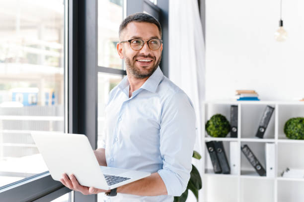 istock Image of office worker man wearing white shirt looking aside, while holding silver laptop during work in business centre 1326385683