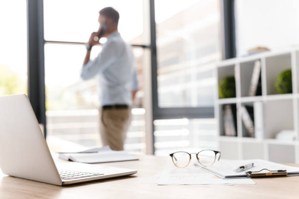 Photo closeup of workplace with office stuff lying on table, while defocused business man speaking on smartphone and looking through big window