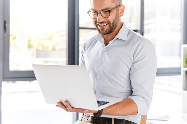 istock Handsome office man wearing white shirt expressing success while holding silver laptop during work in business centre 1326385944