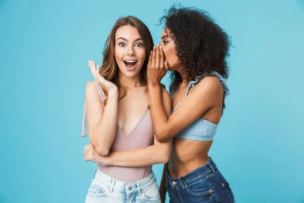 Two cheerful young girls dressed in summer clothes whispering secrets isolated over blue background