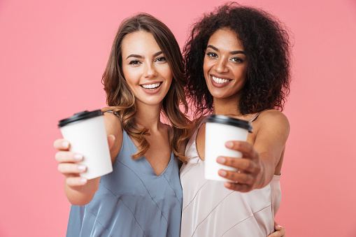 Two beautiful summer girls 20s with different color of skin in dresses smiling at camera and showing paper cups with takeaway coffee isolated over pink background