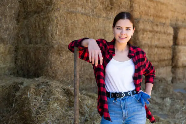 Portrait of a young positive farmer girl standing near haystacks during a work break, holding a pitchfork with gloves
in her hand. Close-up portrait
