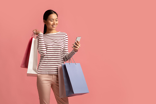 Shopping App. Positive Asian Woman Holding Smartphone And Colorful Paper Shopper Bags, Korean Shopaholic Lady Browsing Mobile Application With Discounts And Sales Info, Standing Over Pink Background