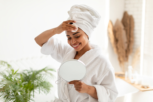 Young Black Lady Removing Makeup With Cotton Pad Looking In Round Mirror Standing In Modern Bathroom At Home. Woman Caring For Face Skin, Beauty And Facial Skincare Concept