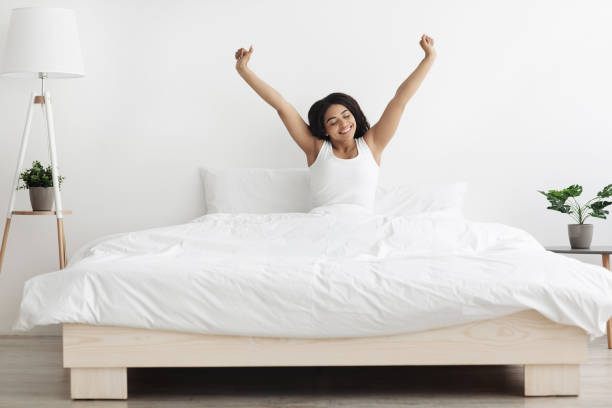 happy african american lady sitting on bed, stretching arms after sleep and enjoying morning in cozy bedroom - wake up stretching women black imagens e fotografias de stock