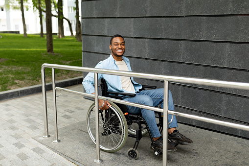 Happy impaired black man in wheelchair entering building on ramp outdoors, full length. Cheerful young guy using disabled-friendly facilities. Handicapped-accessible city concept