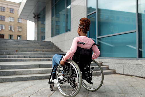 Disability, environment, mobility and social participation concept. Young black impaired lady in wheelchair cannot go up stairs without ramp, feeling limited and unhappy, copy space