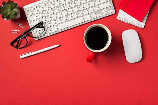 Top view photo of white keyboard mouse flowerpot glasses clips pen red cup of coffee and two notebooks on isolated vivid red background with copyspace