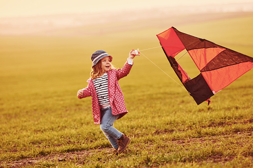 Happy little girl running with kite in hands on the beautiful field at sunrishe time.