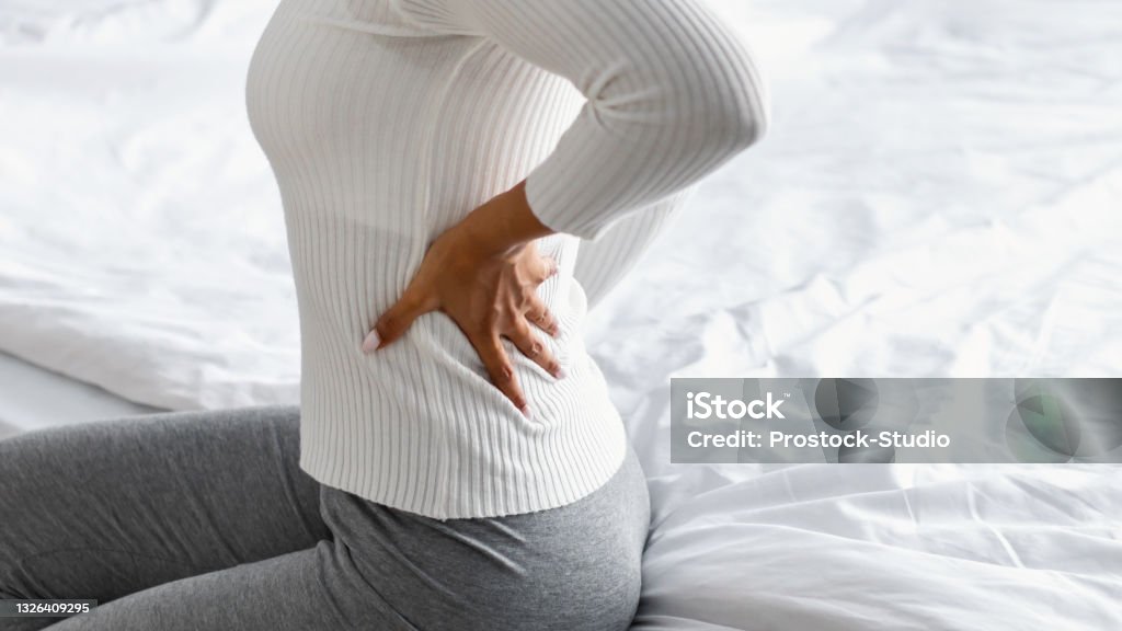 Closeup of black woman with back pain sitting on bed Closeup cropped view of unrecognizable black young woman suffering from pain in body after sleeping, touching massaging her lower back, having uncomfortable bed and mattress, banner, free copy space Backache Stock Photo