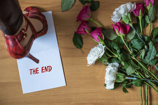 Woman legs in red high heel shoes walking on paper with word the end on a floor with withered flowers. Heart broken concept. Divorce
