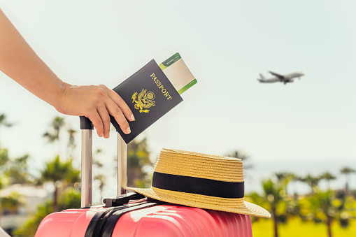 Woman with pink suitcase and amerian passport with boarding pass standing on passengers ladder of airplane opposite sea with palm trees. Tourism concept