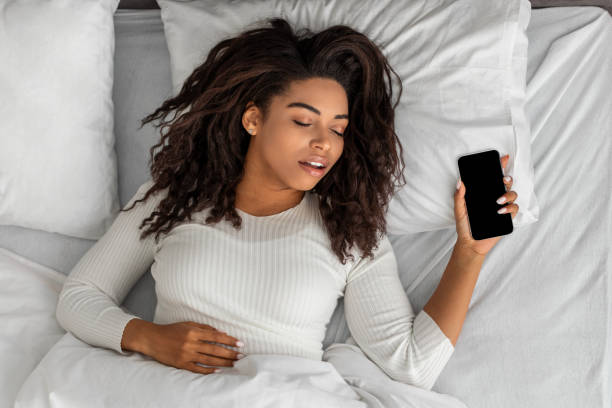 Black woman lying in bed, sleeping and holding mobile phone Asleep African American Woman Sleeping Through Alarm Clock On Phone Lying In Bed In Bedroom At Home In The Morning, Holding Smartphone. Oversleeping, Late For Work, Early Awakening Concept tracker stock pictures, royalty-free photos & images