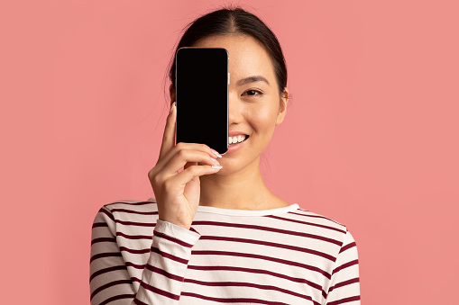 Cheerful Asian Female Covering Face With Blank Black Smartphone, Smiling Young Korean Woman Demonstrating Copy Space For Your Mobile Design And Advertisement, Pink Background, Mockup Image
