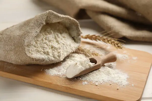 Sack and scoop of flour with wheat ears on wooden board, closeup
