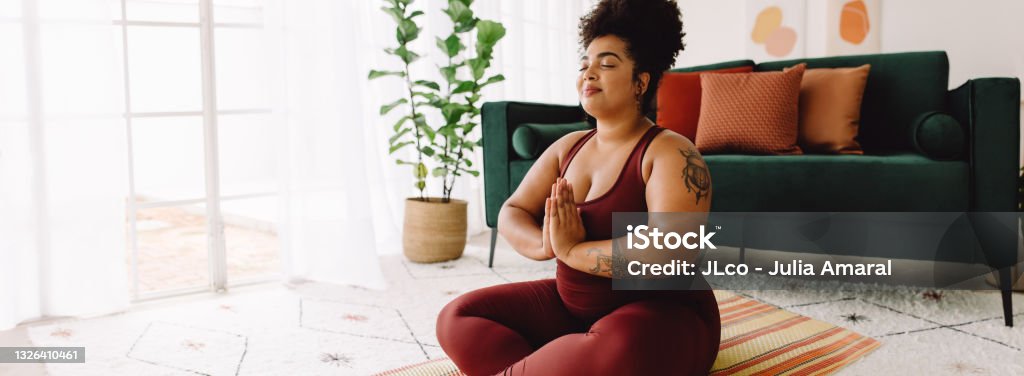 Healthy woman practicing yoga at home Healthy female in practicing meditation yoga at home. Body positive woman exercising with eyes closed and hands joined while sitting cross legged in living room. Relaxation Stock Photo